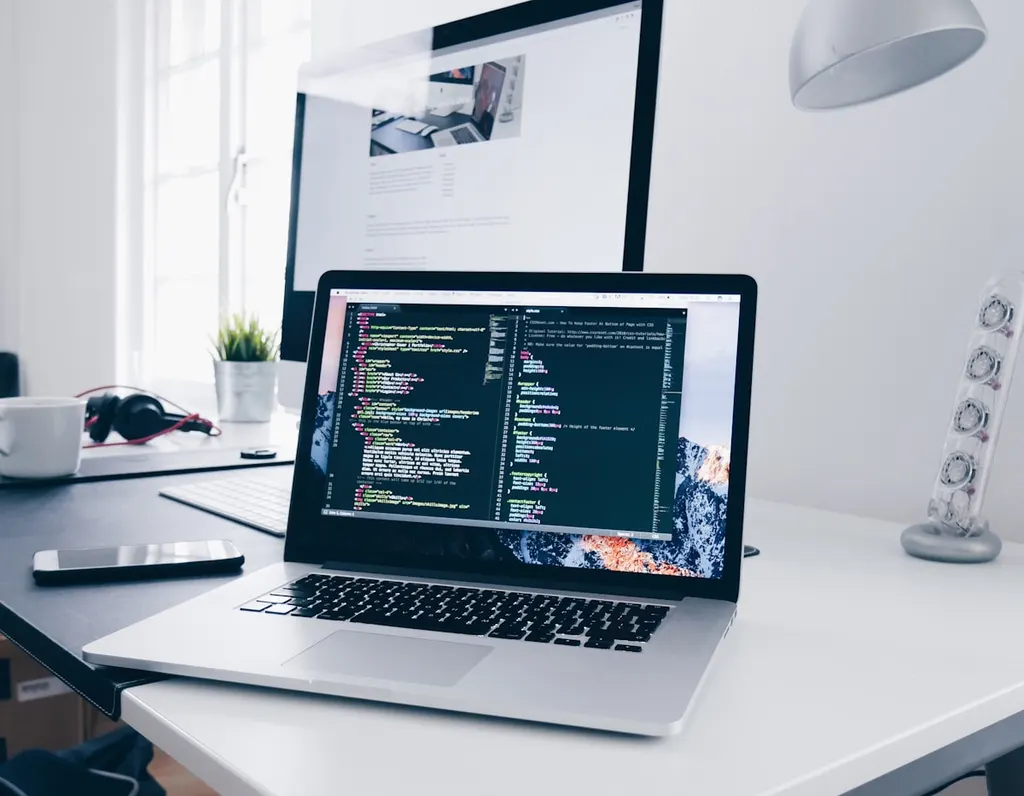 Kod açık bir laptop ve yanında monitör olan çalışma masası